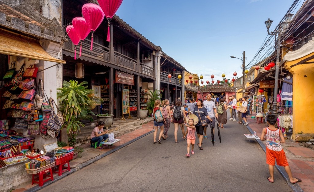 A bustling street in an old town in Vietnam with shops selling colorful textiles and souvenirs, lanterns hanging across the street, and numerous tourists walking and browsing.